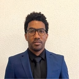 Man with curly hair and glasses in blue suit and black tie against white wall