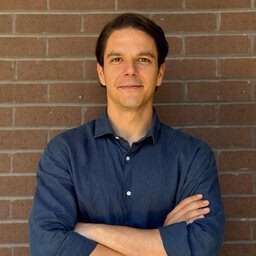 Young man in blue shirt stands with arms crossed against brick wall.