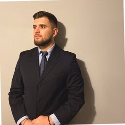 Photograph: bearded man in navy suit and tie looking sideways against a gray wall